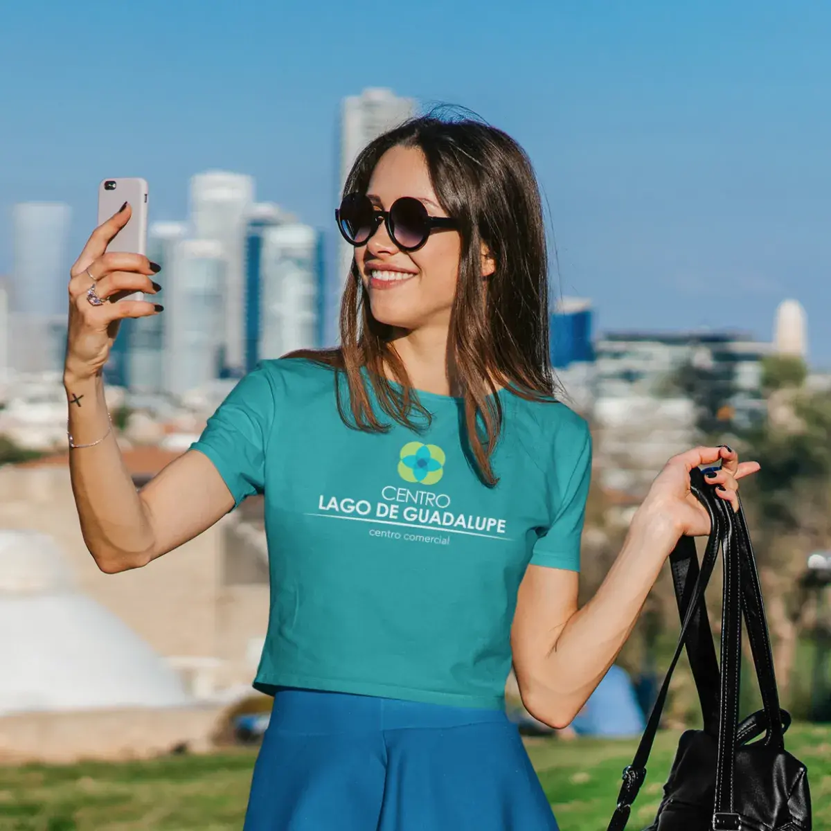 Mujer sonriente con lentes de sol tomándose una selfie con la silueta urbana de fondo en Centro Lago de Guadalupe.